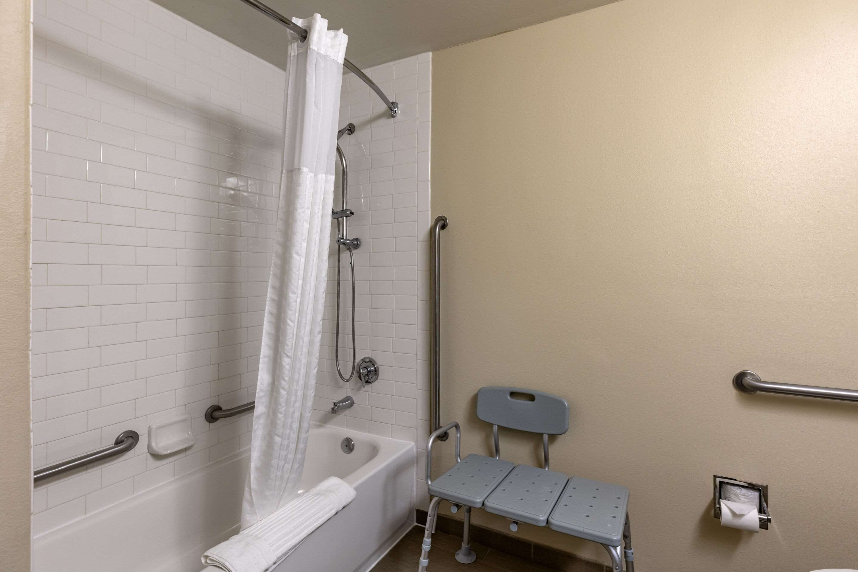 A bathroom with a white-tiled shower, a white bathtub, a grab bar, a wheelchair-accessible bench, and a hand dryer.