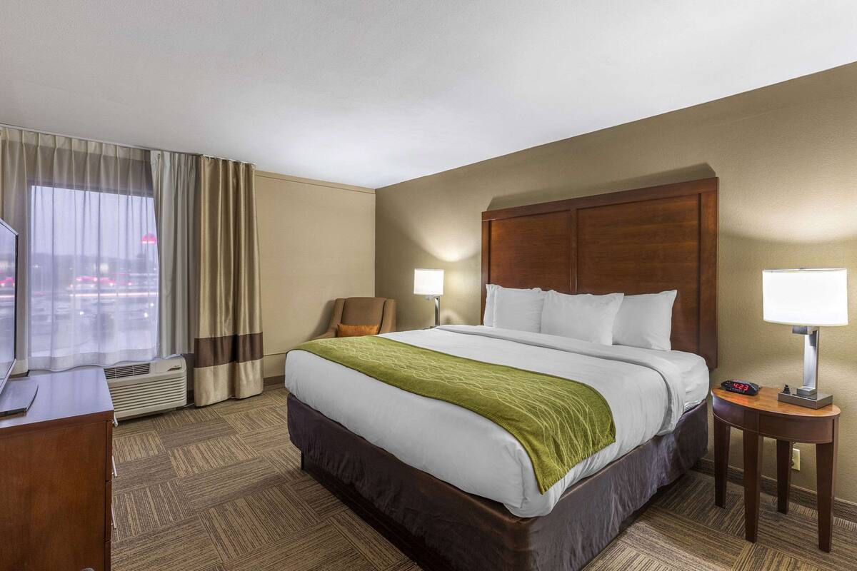 A hotel room with a large bed, two bedside lamps, a chair, a desk, and a window with curtains.