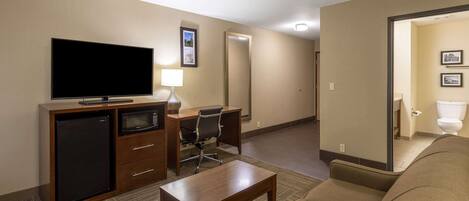 A hotel room with a flat-screen TV, a mini-fridge, a microwave, a desk, a chair, a sofa, and a bathroom visible through an open door.