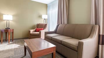 Ett hotellrum med en beige soffa, ett träbord, två lampor och ett fönster med gardiner.