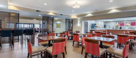 A spacious dining area with multiple tables and chairs, a kitchen area in the background, and a modern ceiling design.