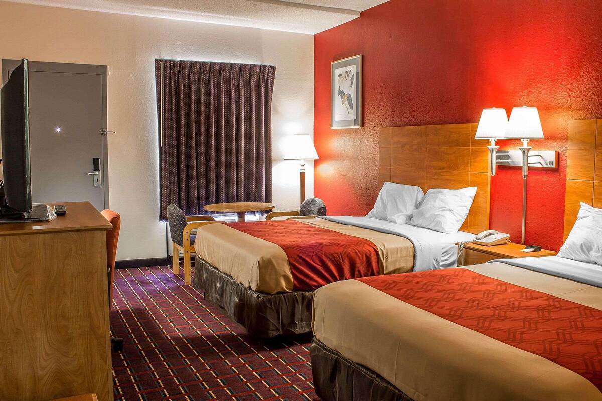 Ein Hotelzimmer mit zwei Betten, einem Schreibtisch, einem Stuhl, einem kleinen Tisch, einer Lampe und einem Bild an der Wand.