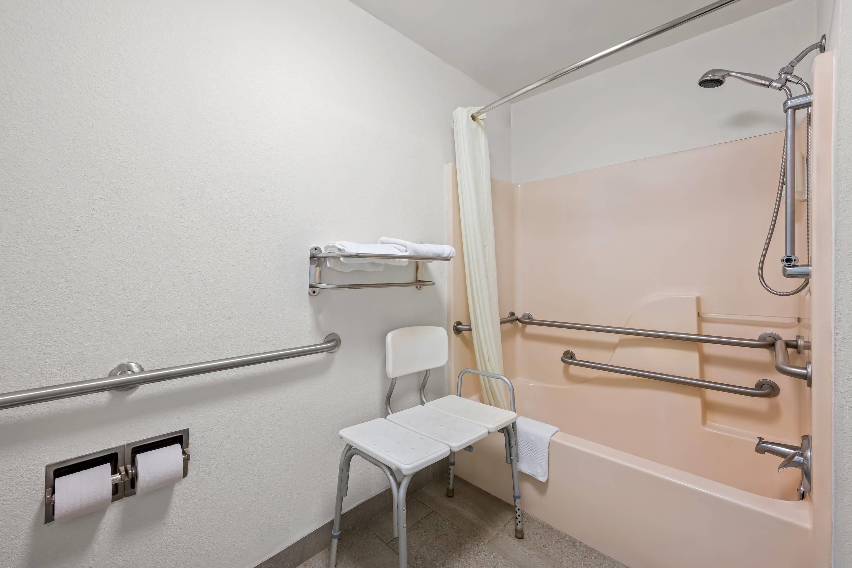 Ein Badezimmer mit barrierefreier Dusche, Haltegriffen, einer Toilette mit Papierhandtuchspender und einem weißen Stuhl.