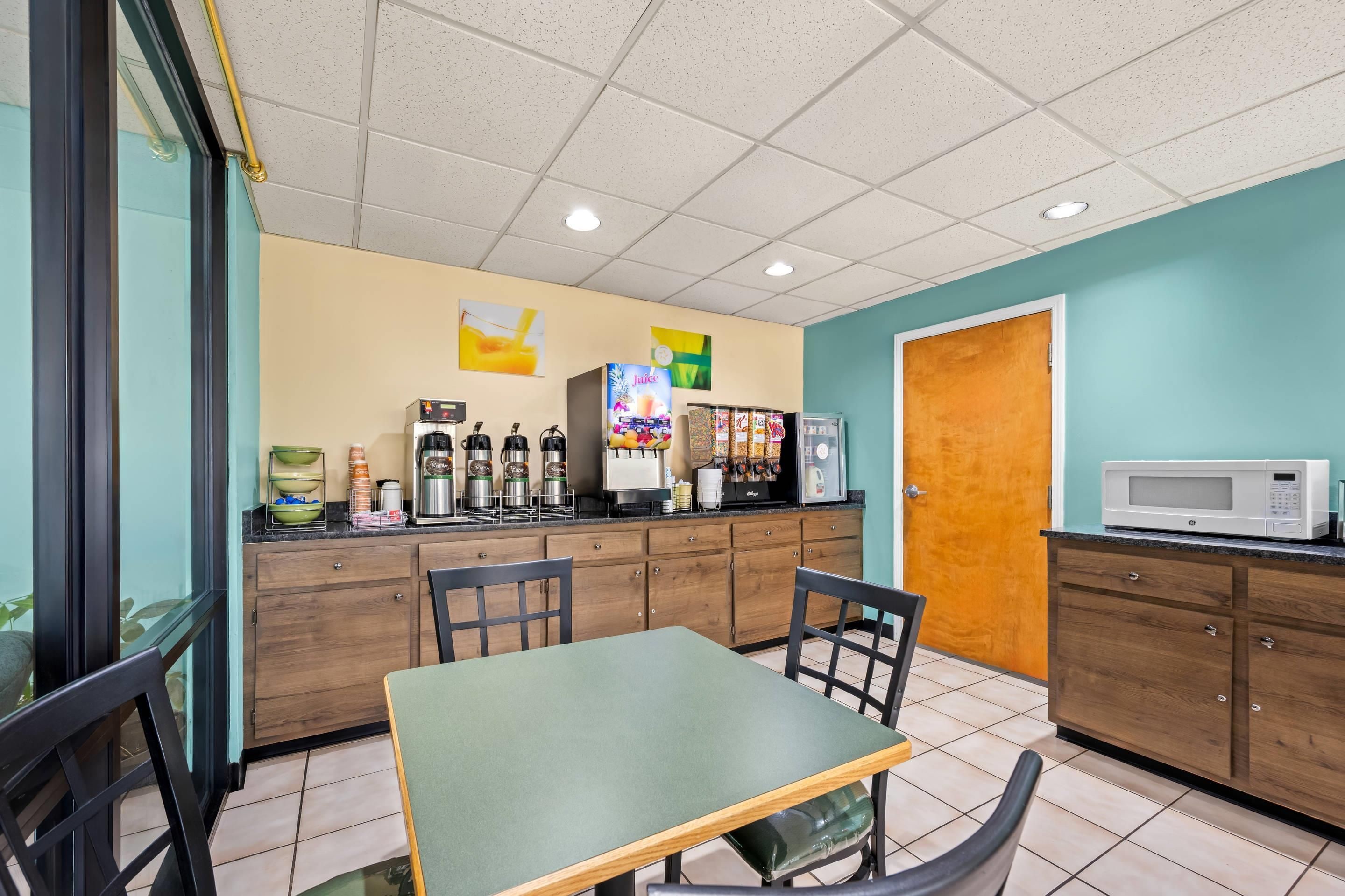 A small cafe with a green table, chairs, a coffee machine, a refrigerator, and a microwave.