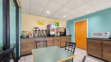 A small cafe with a green table, chairs, a coffee machine, a refrigerator, and a microwave.