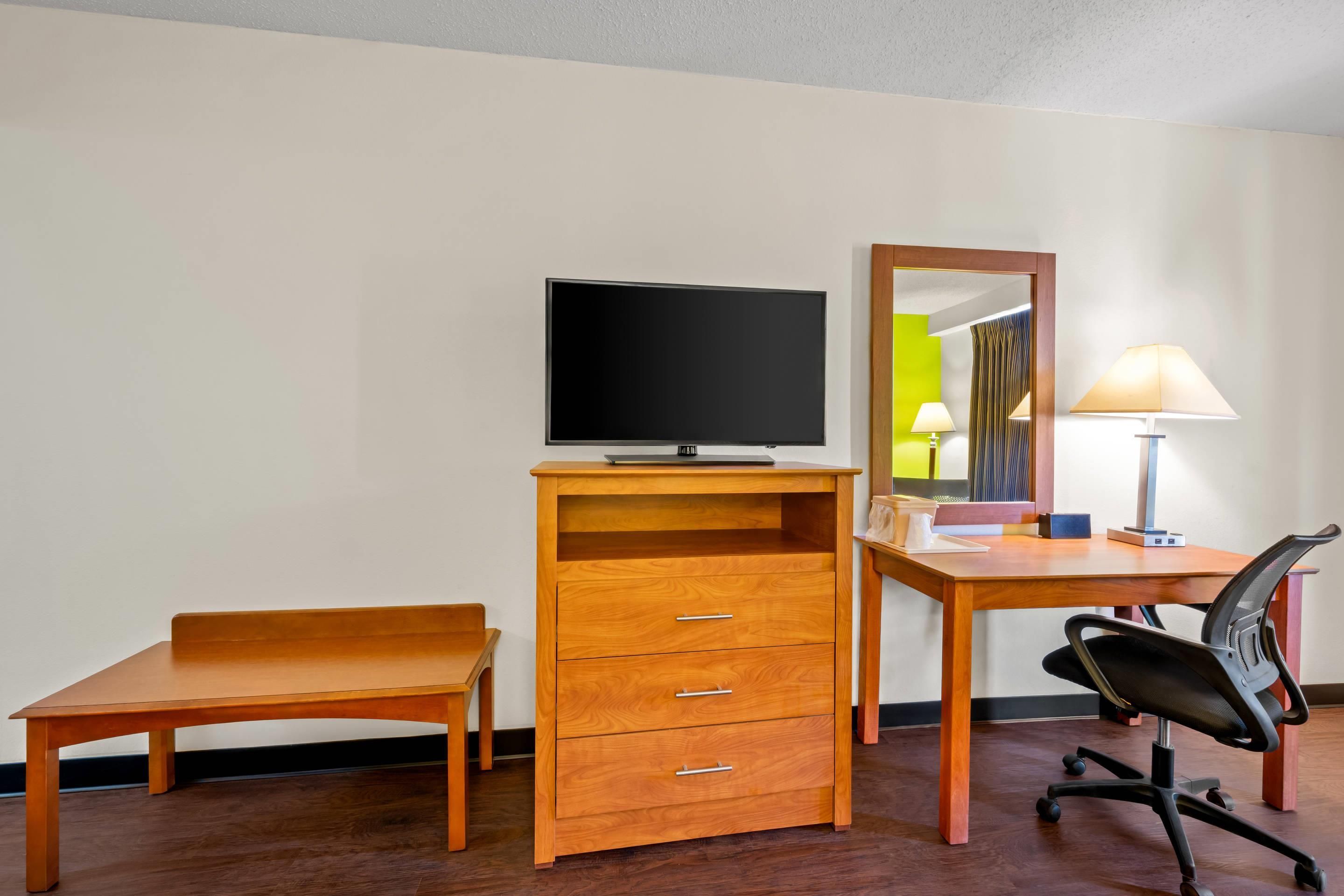 Una habitación de hotel con un escritorio de madera, una silla, un televisor, un espejo y una lámpara.