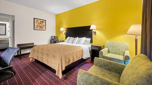 A hotel room with a bed, two bedside tables, a chair, a desk, a lamp, a television, and a patterned carpet. - Quality Inn Henderson I-85 (Henderson)