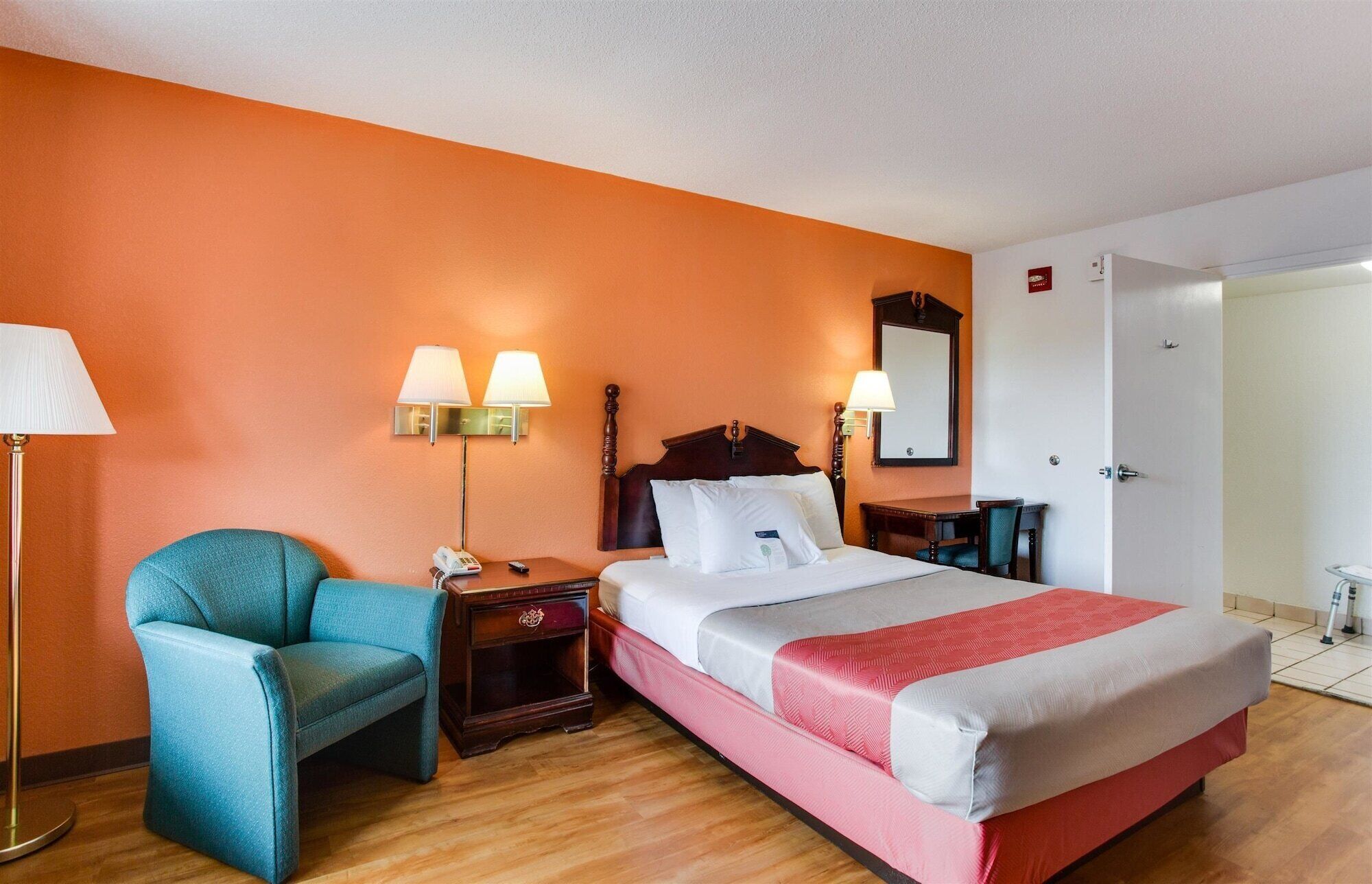 A hotel room with a bed, a chair, a nightstand, a lamp, and a mirror.