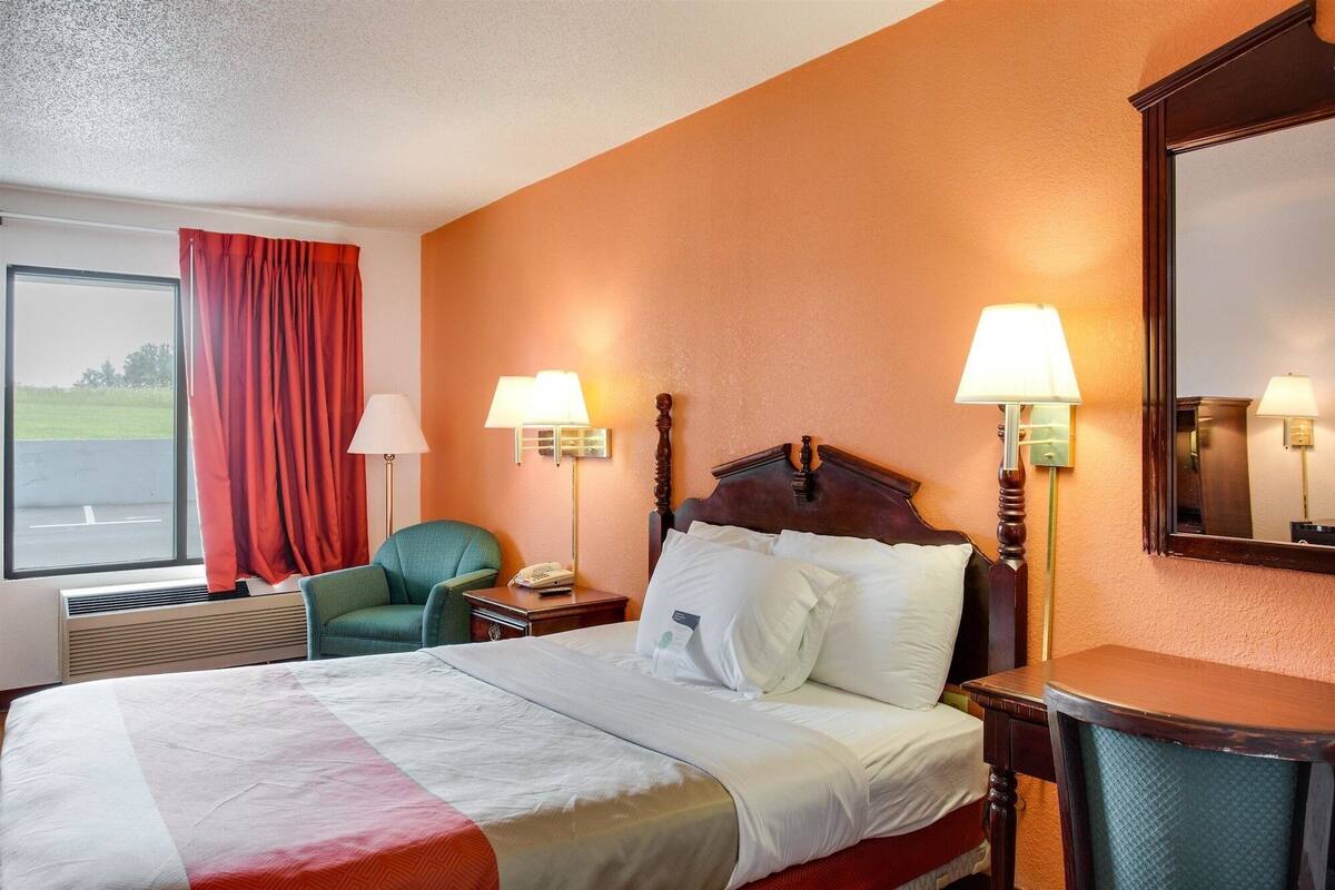 A hotel room with a large bed, two bedside lamps, a chair, a desk, and a window with red curtains.