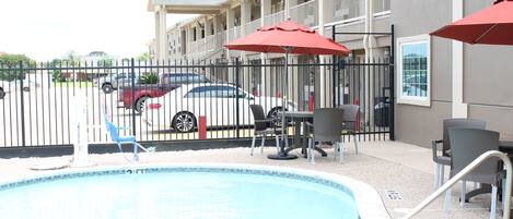 A swimming pool with a tiled edge, outdoor seating with red umbrellas, and a building with balconies in the background.