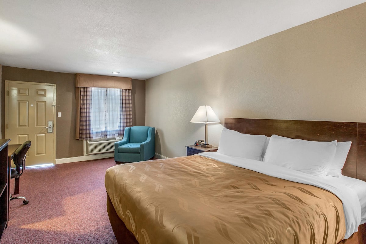 A hotel room with a large bed, a desk, a chair, a lamp, and a window with curtains.