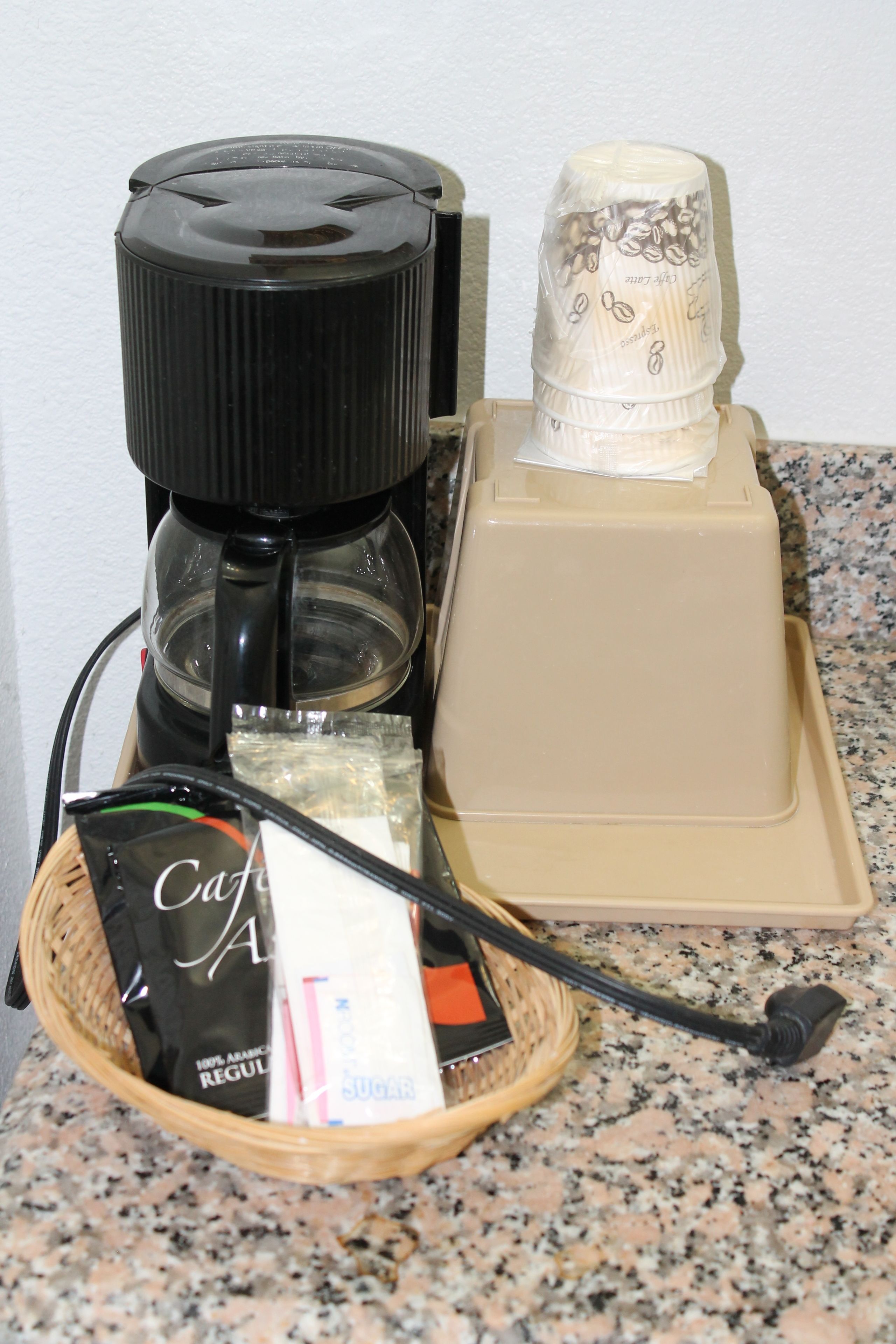 A coffee maker, a coffee pot, a basket with coffee packets, and a spoon on a granite countertop.