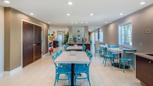 Uma sala de jantar espaçosa com uma mesa central rodeada por cadeiras azuis, uma área de cozinha com balcão de refeições rápidas e uma grande janela que proporciona luz natural.