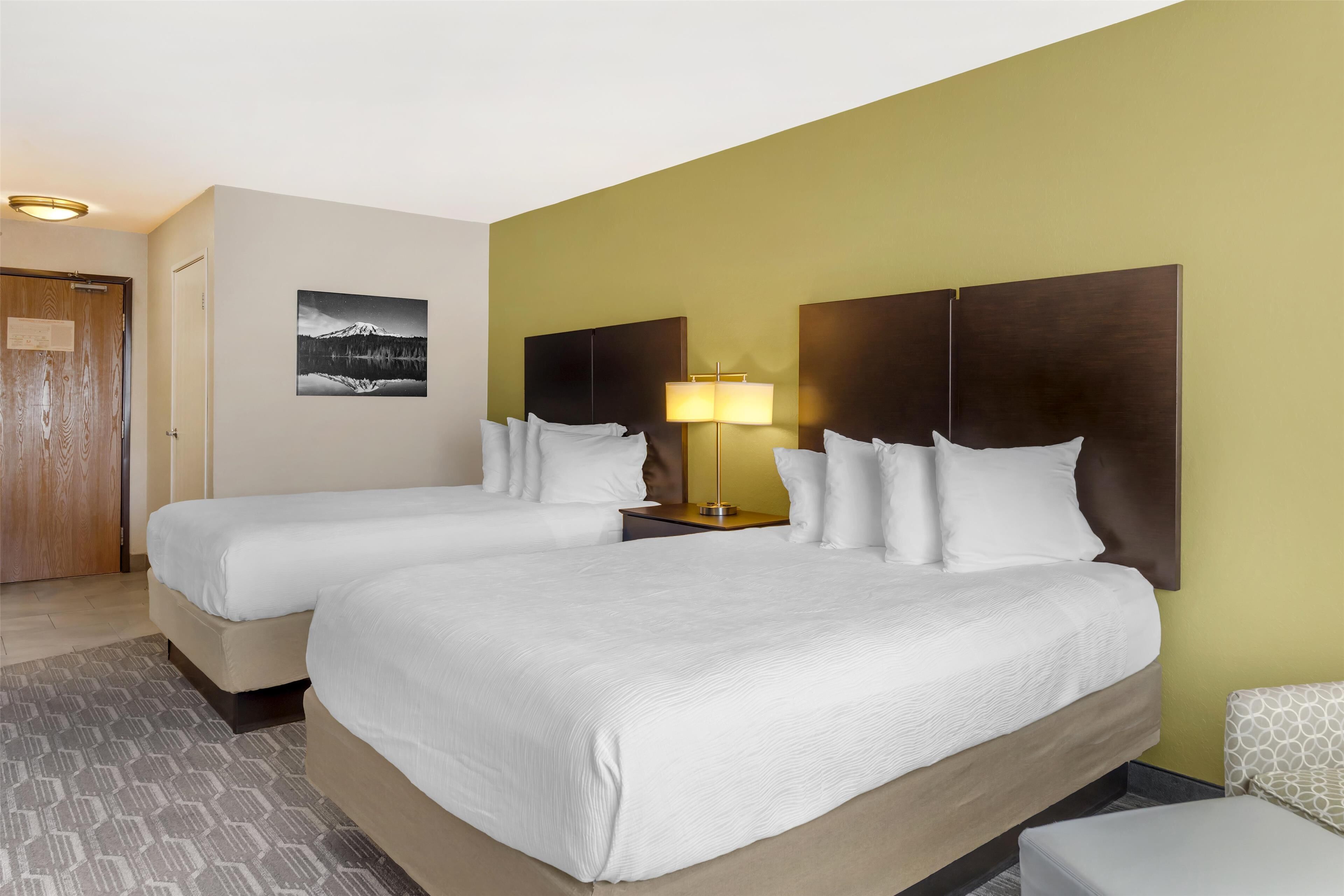 A hotel room with two beds, a nightstand with a lamp, a wall-mounted picture, and a patterned rug.