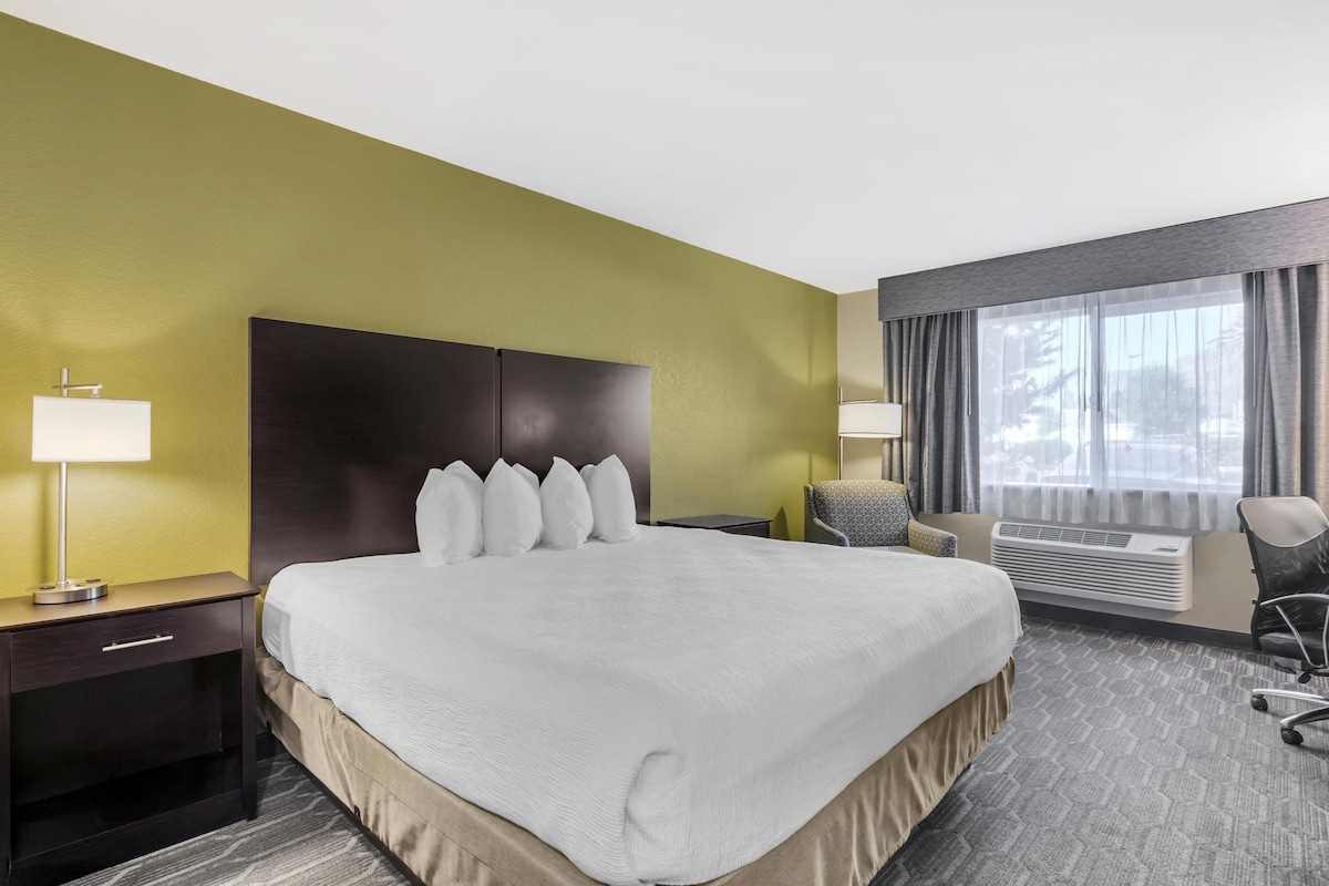 A hotel room with a large bed, a bedside table with a lamp, a chair, and a window with curtains.