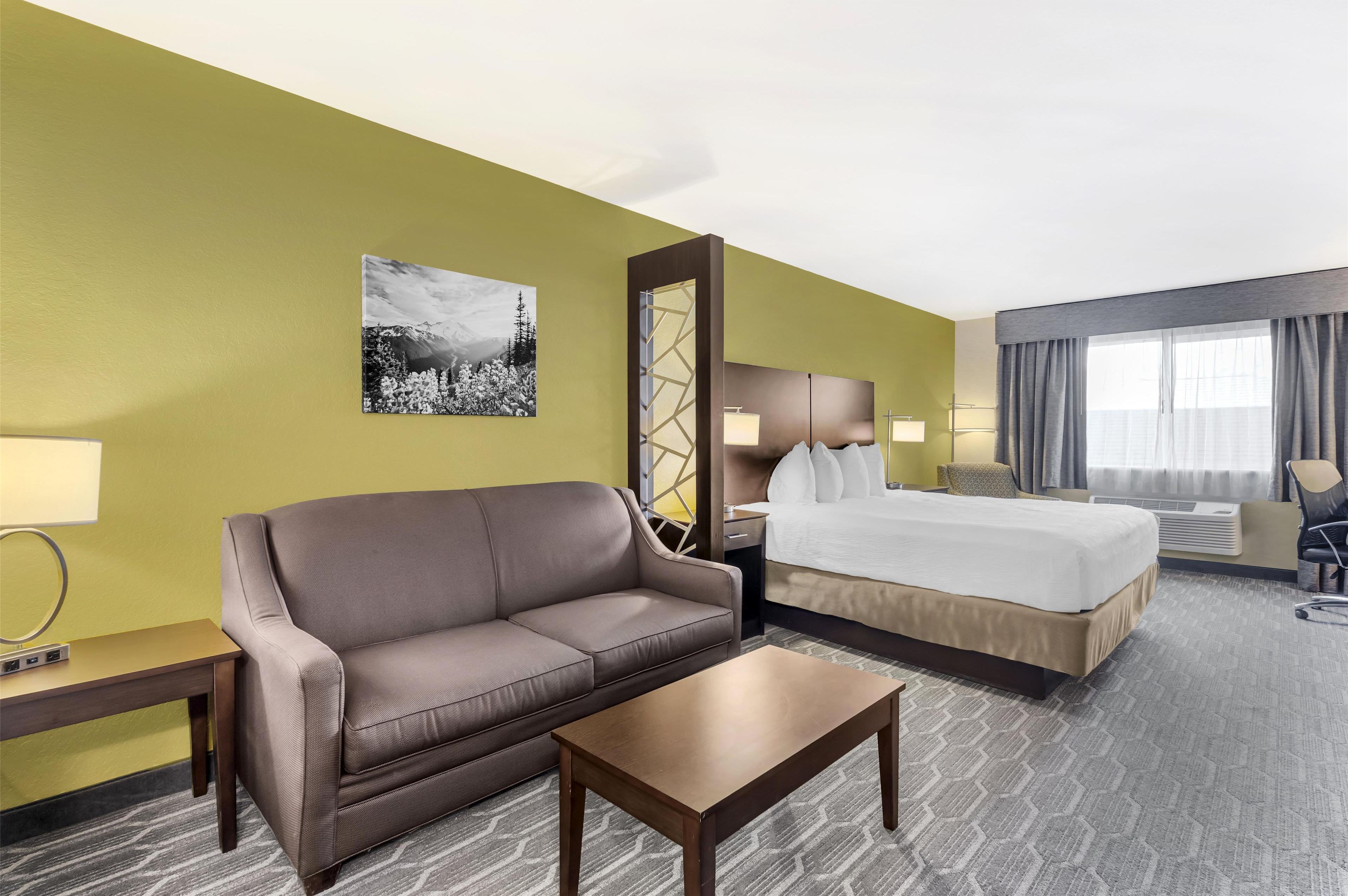 A hotel room with a brown sofa, a bed, a small table, a nightstand, a lamp, a mirror, and a picture on the wall.