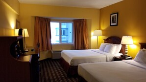 A hotel room with two beds, a desk with a lamp, a TV, and a window with curtains. - The Comstock Inn and Conference Center (Owosso)