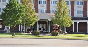 A multi-story building with a central entrance, surrounded by trees and a parking area. - The Comstock Inn and Conference Center (Owosso)