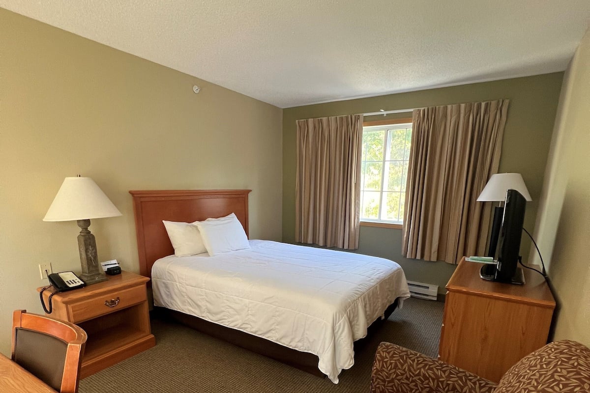 A hotel room with a bed, a desk, a chair, a nightstand with a lamp, a TV, and a window with curtains.