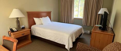 A hotel room with a bed, a desk, a chair, a nightstand with a lamp, a TV, and a window with curtains.