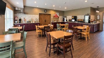 A spacious dining area with multiple wooden tables and chairs, a kitchen with modern appliances, and a brick wall.