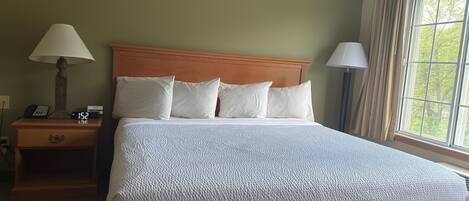 A hotel room with a large bed, a wooden headboard, two bedside lamps, a nightstand with a phone and a digital clock, and a window with curtains.