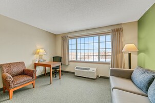 Quarto de hotel com uma poltrona estampada, um sof á, uma mesa com abajur, uma cadeira, uma janela grande com cortinas e um aparelho de ar condicionado.