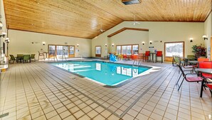 Una piscina interior con suelo de baldosas, techo de madera y zonas de estar con mesas y sillas.