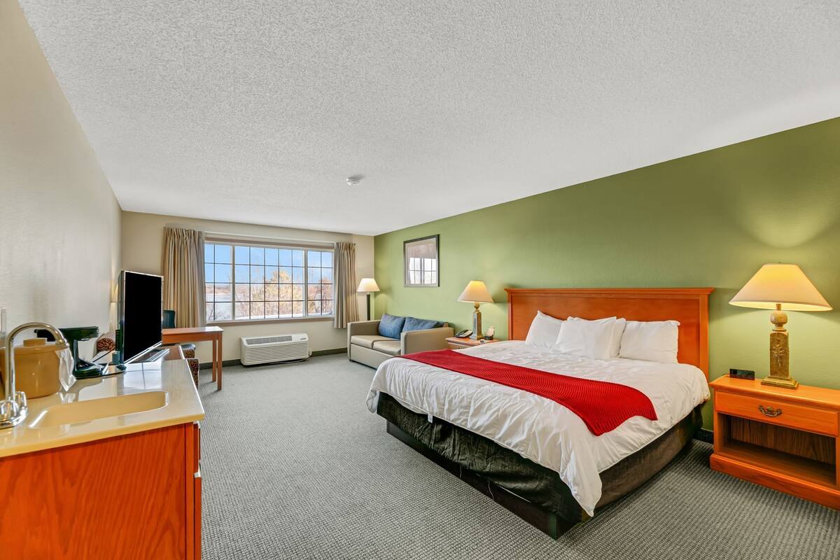 A hotel room with a bed, a desk with a sink, a chair, a window with curtains, and a lamp.