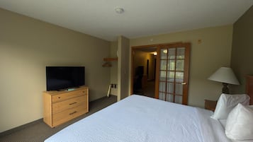 A hotel room with a large bed, a wooden dresser with a TV, a lamp, and a door leading to another room.
