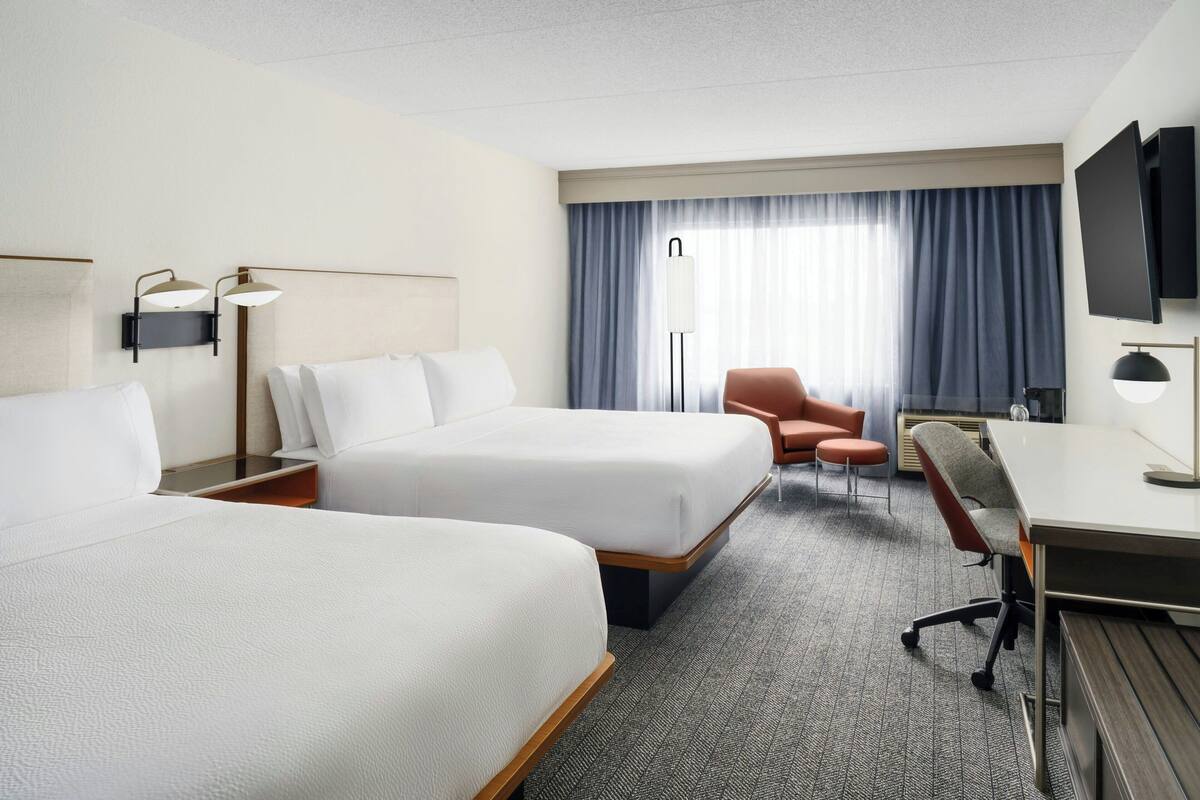 Ein Hotelzimmer mit zwei Betten, einem Schreibtisch, einem Stuhl, einem Fernseher und einem Fenster mit Vorhängen.
