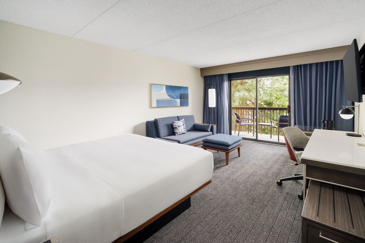 A hotel room with a bed, a sofa, a desk, and a view of the outdoors through a sliding glass door.