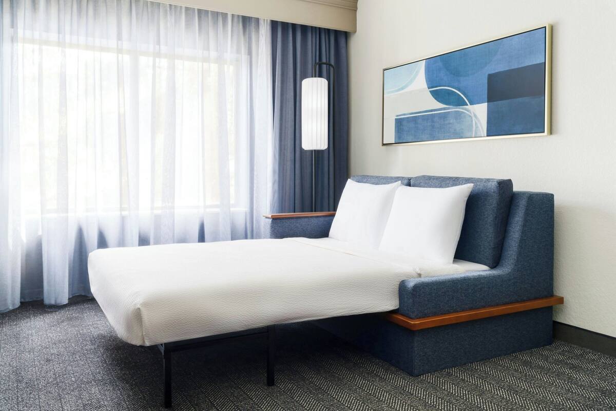 Ein modernes Hotelzimmer mit einem Bett, einem großen Fenster mit transparenten Vorhängen, einer Stehlampe und einem abstrakten Kunstwerk an der Wand.