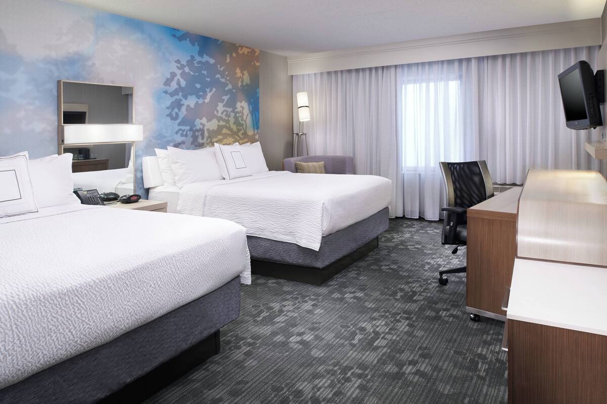 Ein Hotelzimmer mit zwei Betten, einem Schreibtisch, einem Stuhl, einem Fernseher und einem großen Fenster mit Vorhängen.
