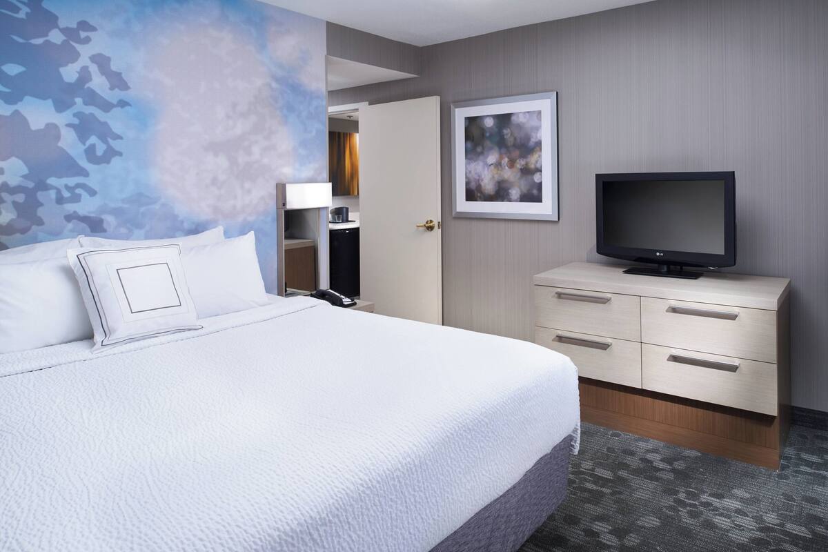 Ein Hotelzimmer mit einem großen Bett, einem Fernseher auf einer Kommode, einer Tür zu einem Badezimmer und einer Wand mit Wandgemälde von Wolken.