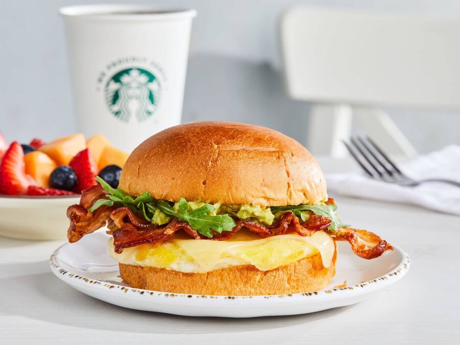 Un sandwich au bacon, à la laitue et au fromage servi dans un pain, une assiette de fruits et, au fond, une tasse Starbucks.
