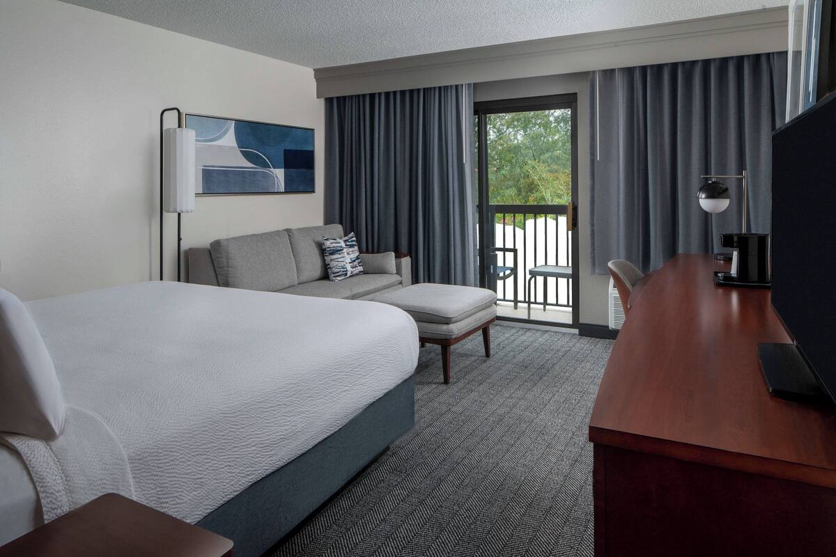 A hotel room with a large bed, a sofa, a desk with a TV, and a balcony with curtains.