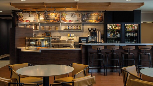 A bar area with stools and a counter, a dining area with round tables and chairs, and shelves with bottles and a coffee machine. - Courtyard by Marriott New Orleans Covington/Mandeville (Covington)