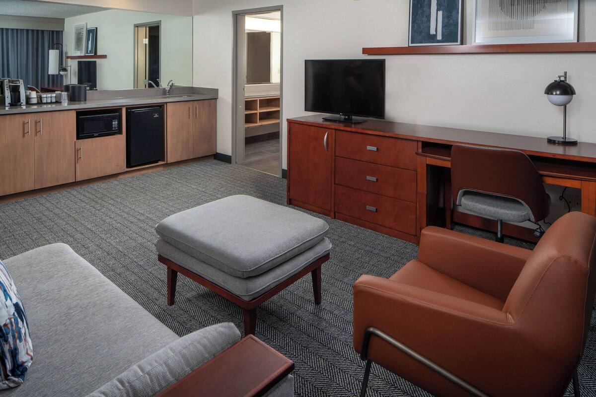 A hotel room with a kitchenette, a TV, a desk, a chair, and a sofa.