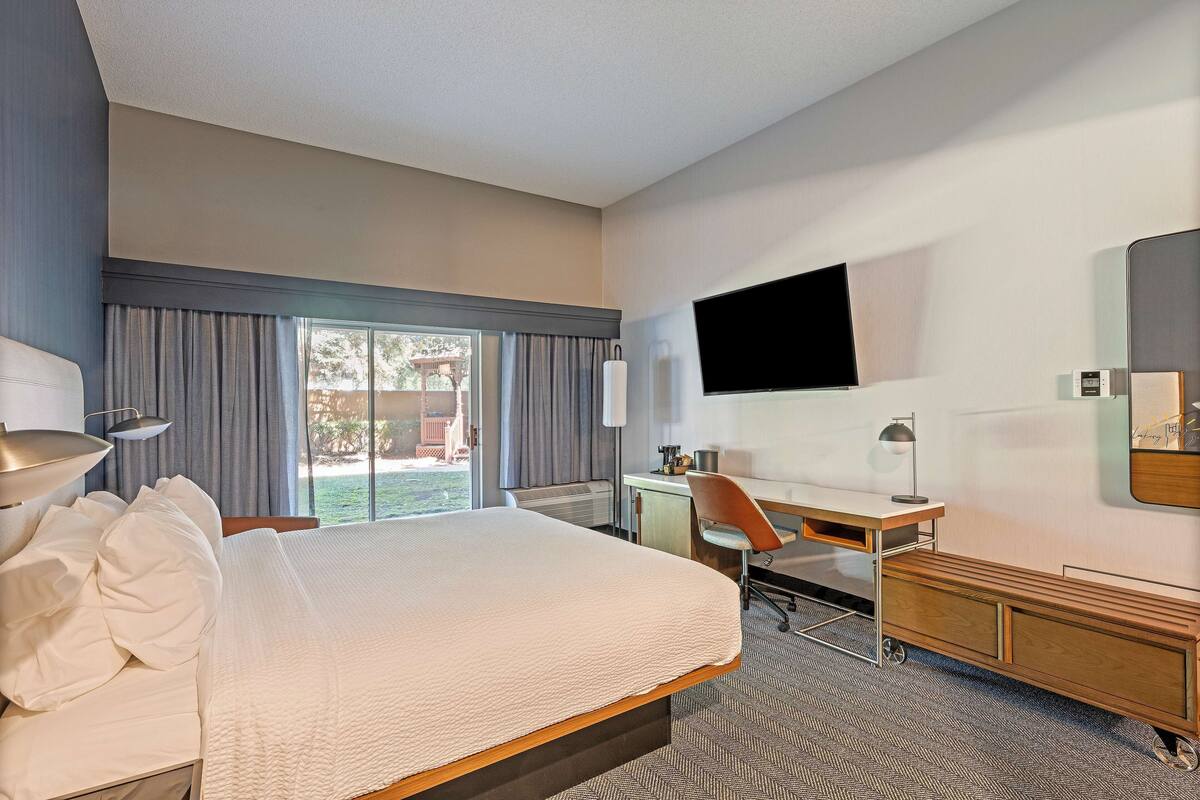 A hotel room with a large bed, a desk, a chair, a TV mounted on the wall, and a window with curtains.