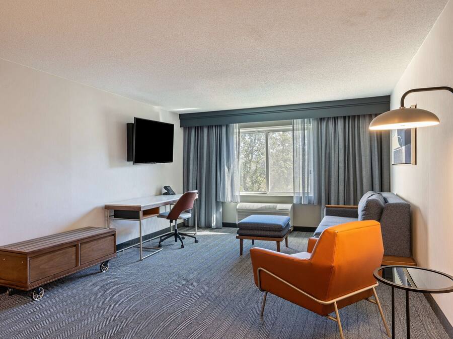 A hotel room with a desk, chair, sofa, ottoman, and a TV mounted on the wall.