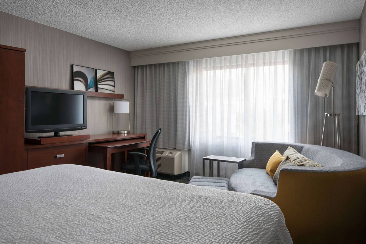 A hotel room with a bed, a desk with a TV, a chair, a small table, and a lamp.