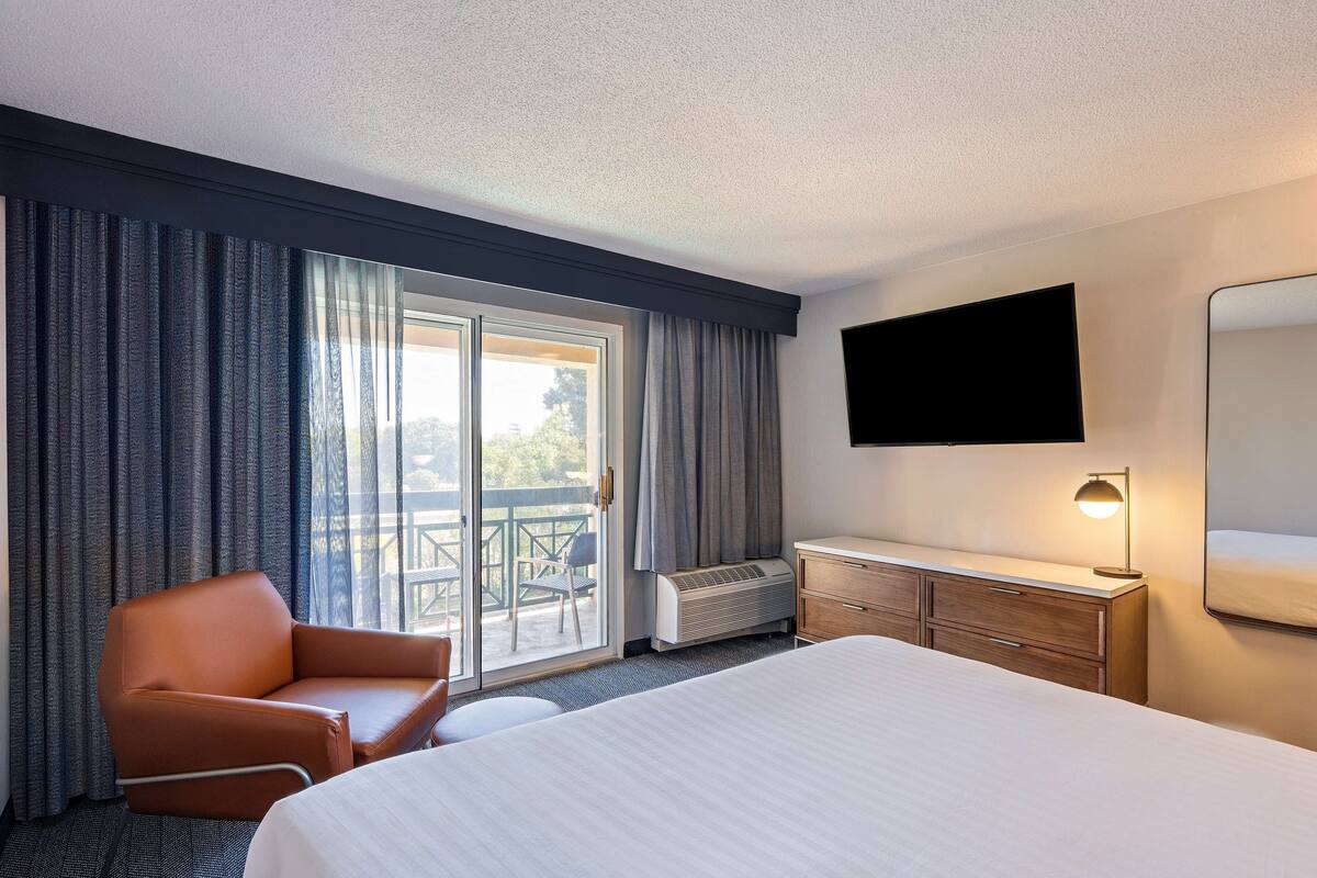 A hotel room with a bed, a desk, a chair, a TV, and a sliding door to a balcony.