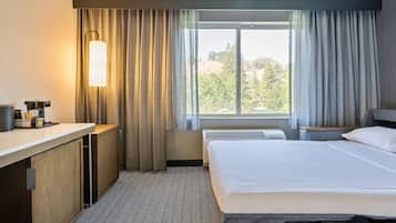 Ein Hotelzimmer mit einem Bett, einem Schreibtisch mit Lampe, einem Fenster mit Vorhängen und Blick auf Bäume.