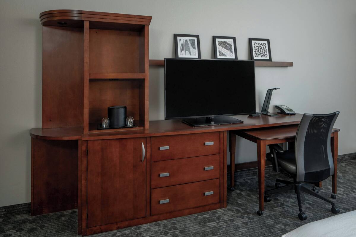 A wooden desk with a flat-screen TV, a lamp, and a chair. There are three drawers and a shelf with pictures.