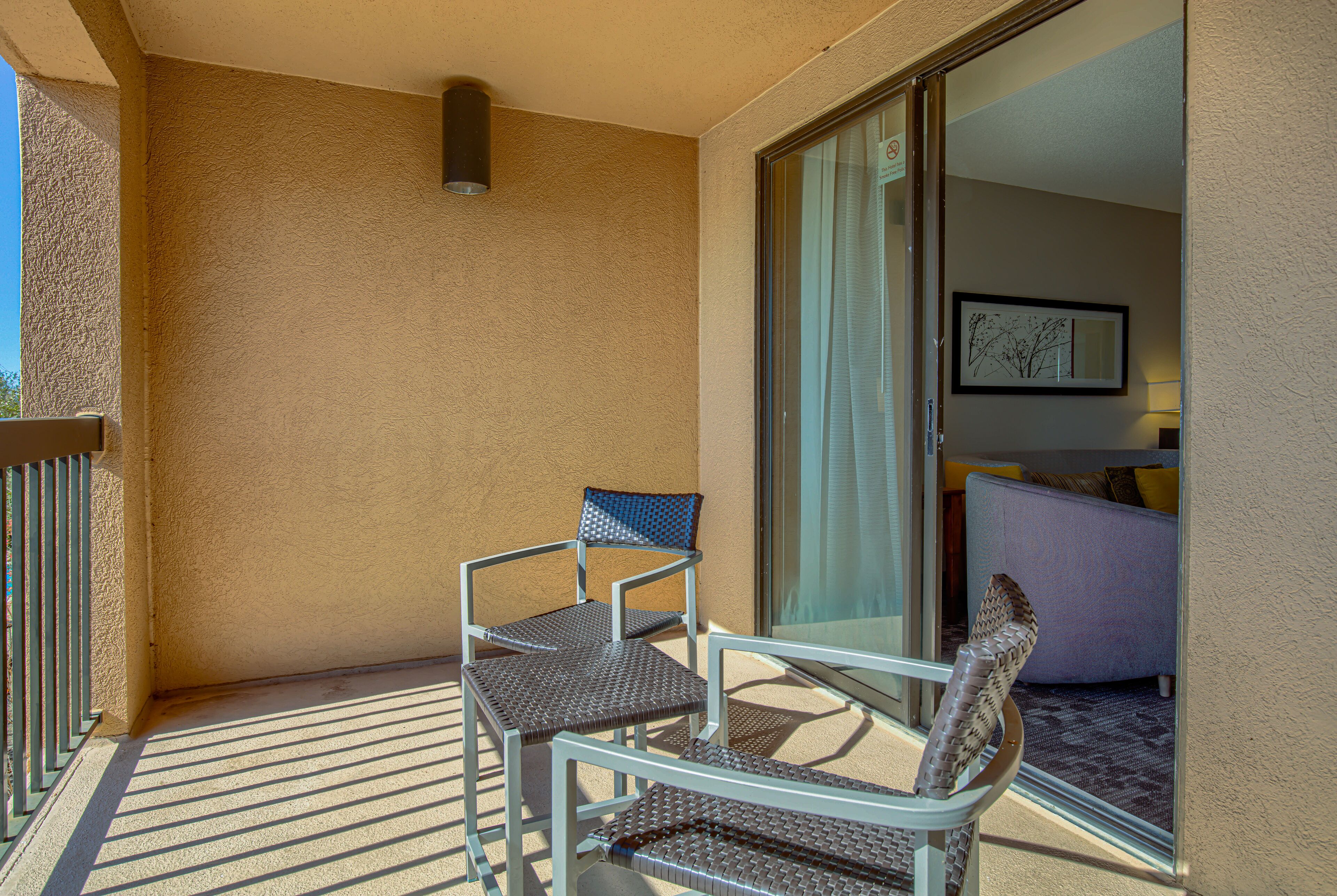 Un balcon avec deux fauteuils en osier et une porte vitrée donnant sur une pièce intérieure, où se trouvent un canapé et un tableau encadré accroché au mur.