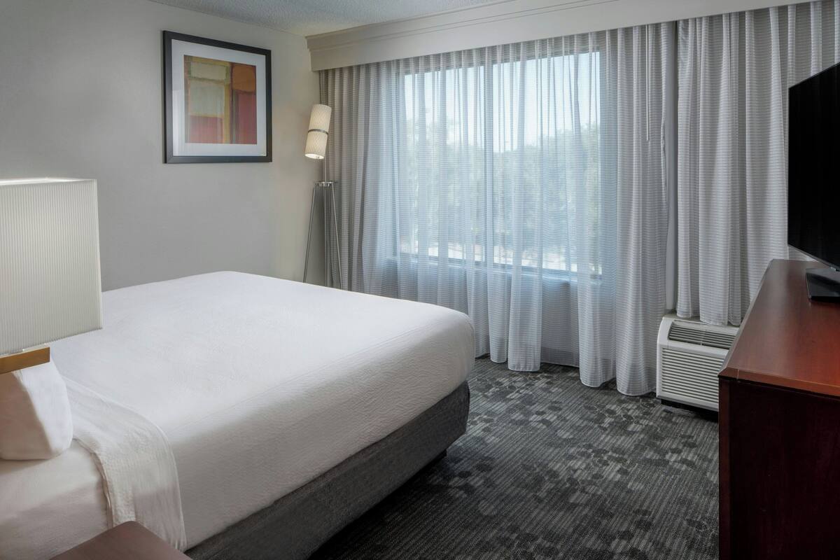 A hotel room with a bed, a desk, a lamp, a picture on the wall, and a window with curtains.