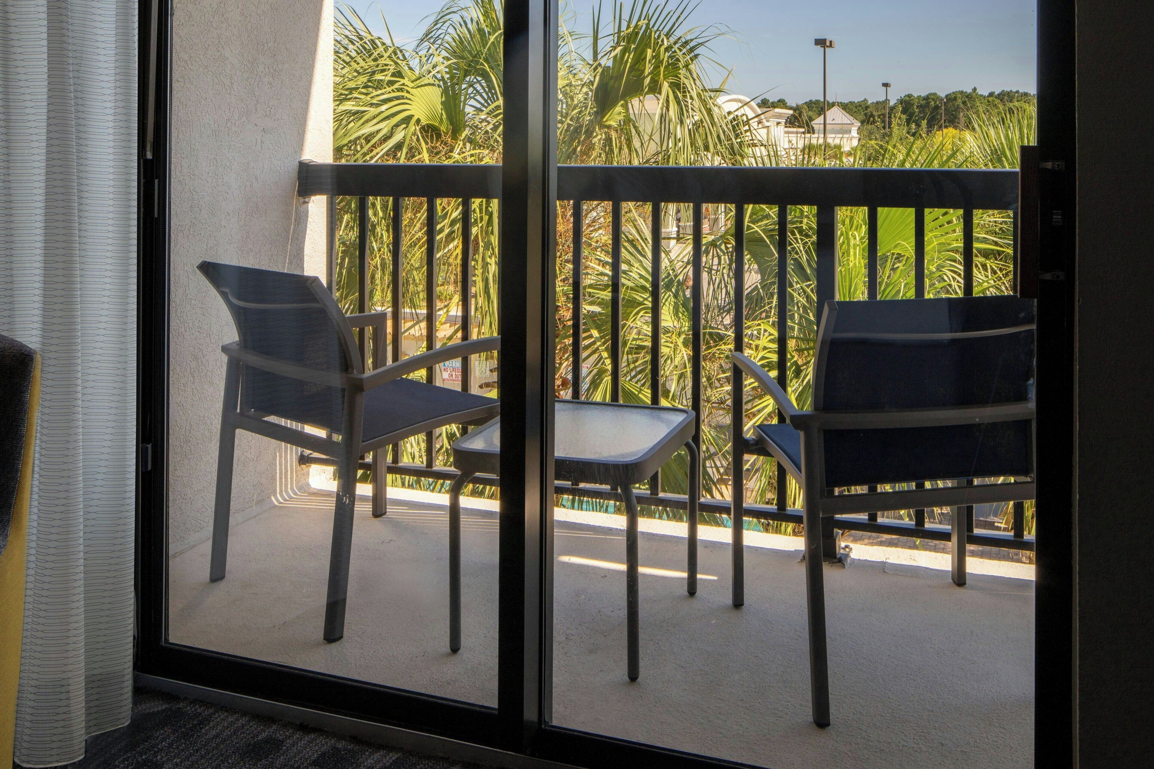 A balcony with two chairs and a table, overlooking a garden with palm trees and a building in the background.