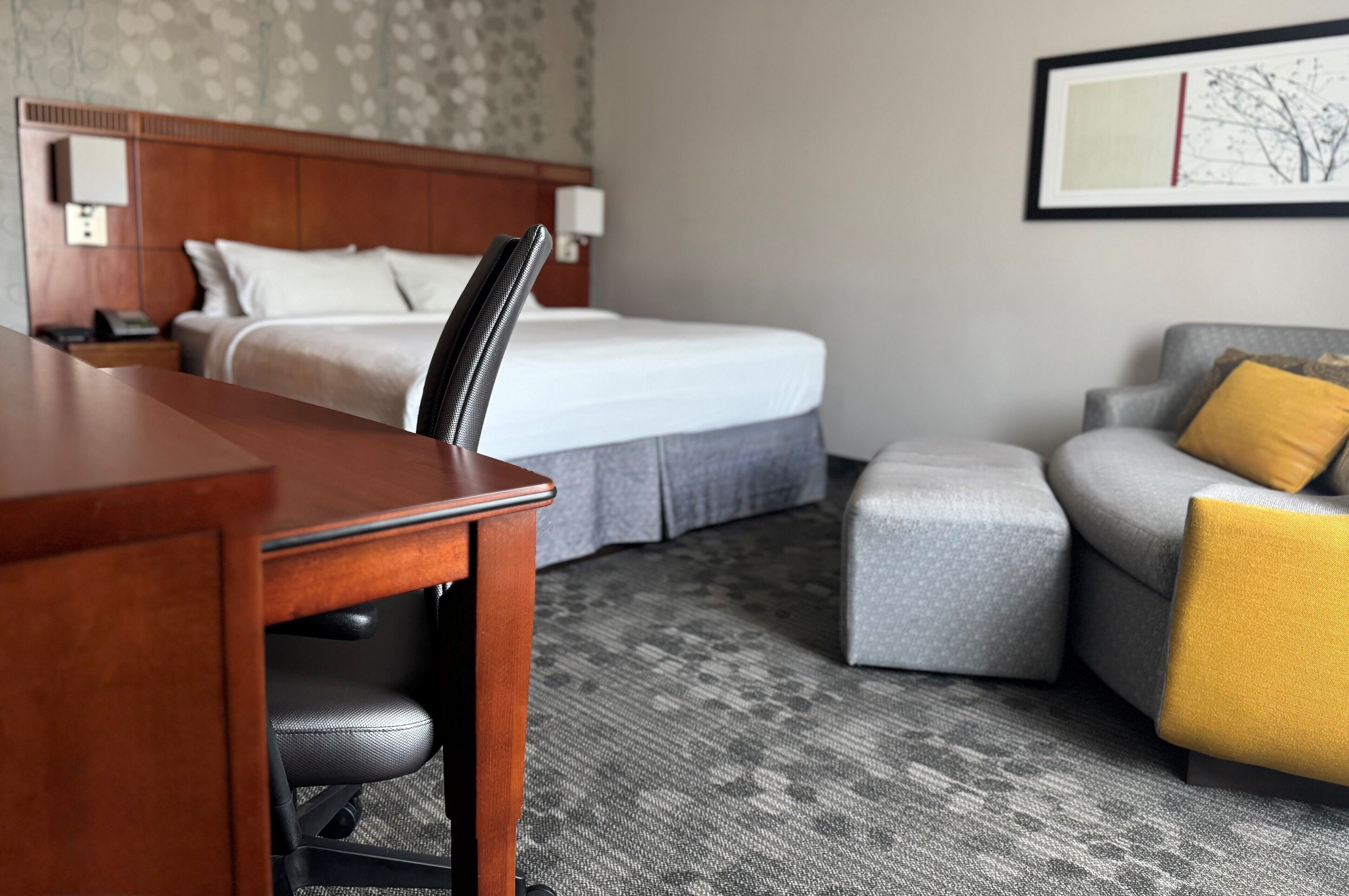 A hotel room with a wooden desk, a grey sofa, a bed with white linens, and a framed picture on the wall.