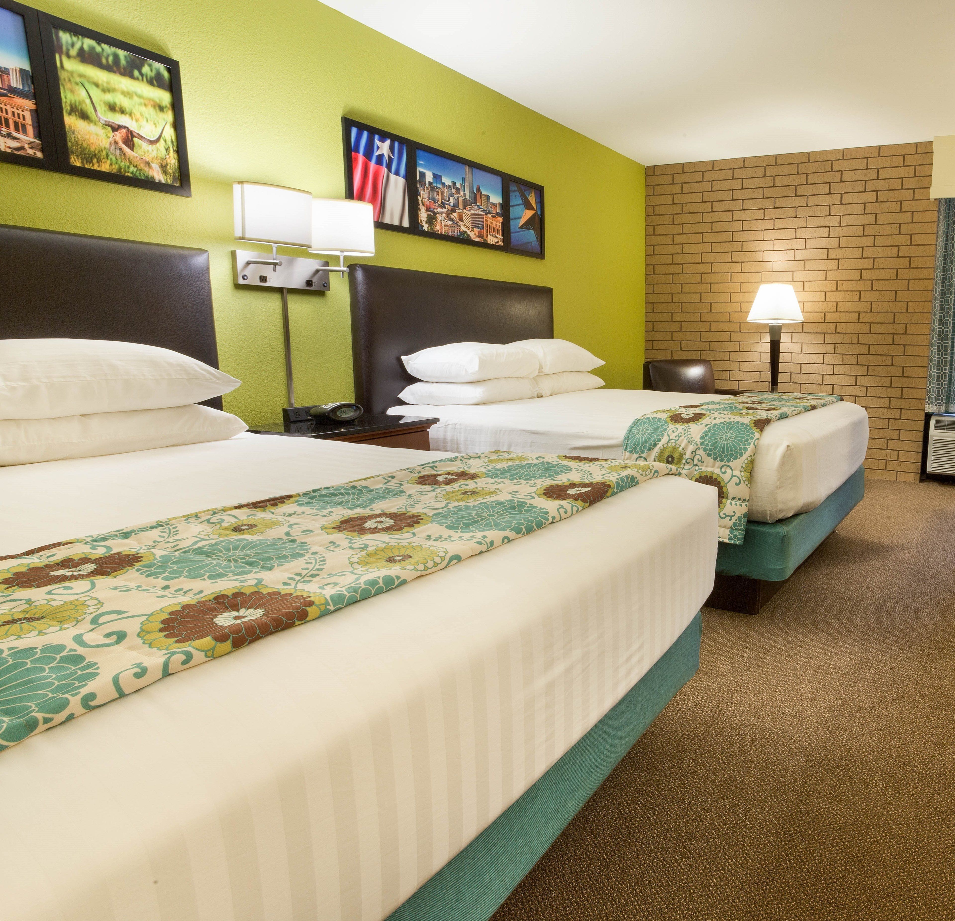 Habitación de hotel con dos camas, una mesita de noche con lámpara y una pared de ladrillo.
